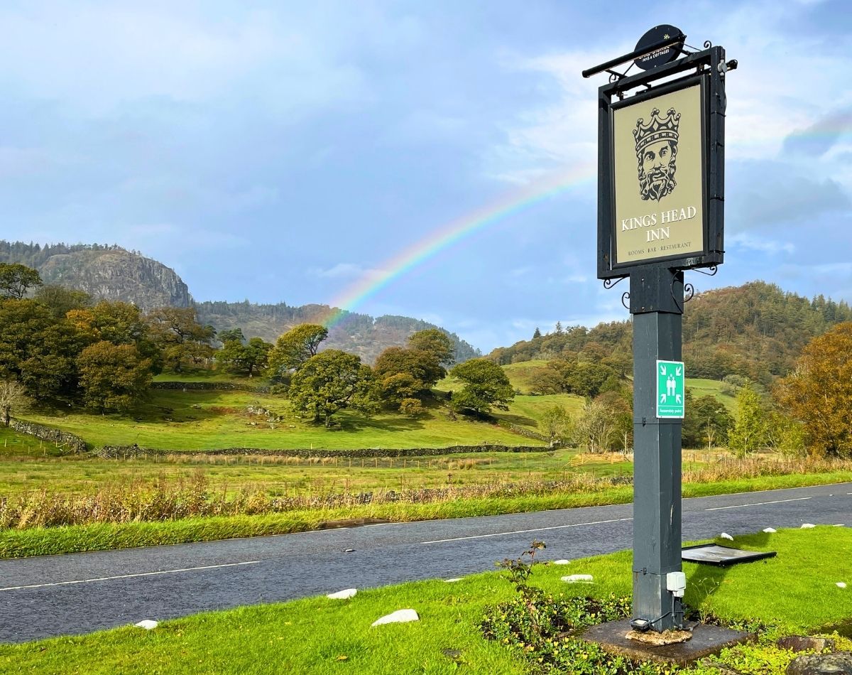 The Kings Head Inn pub sign standing by the A591 road near Thirlmere, with a bright rainbow arching over the Lake District fells in the background.