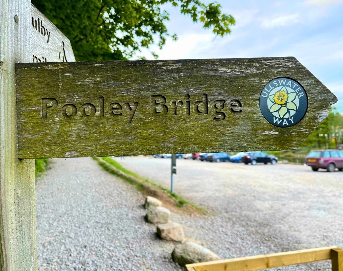 arved wooden signpost pointing to Pooley Bridge with a yellow daffodil Ullswater Way marker attached, set against a gravel car park.