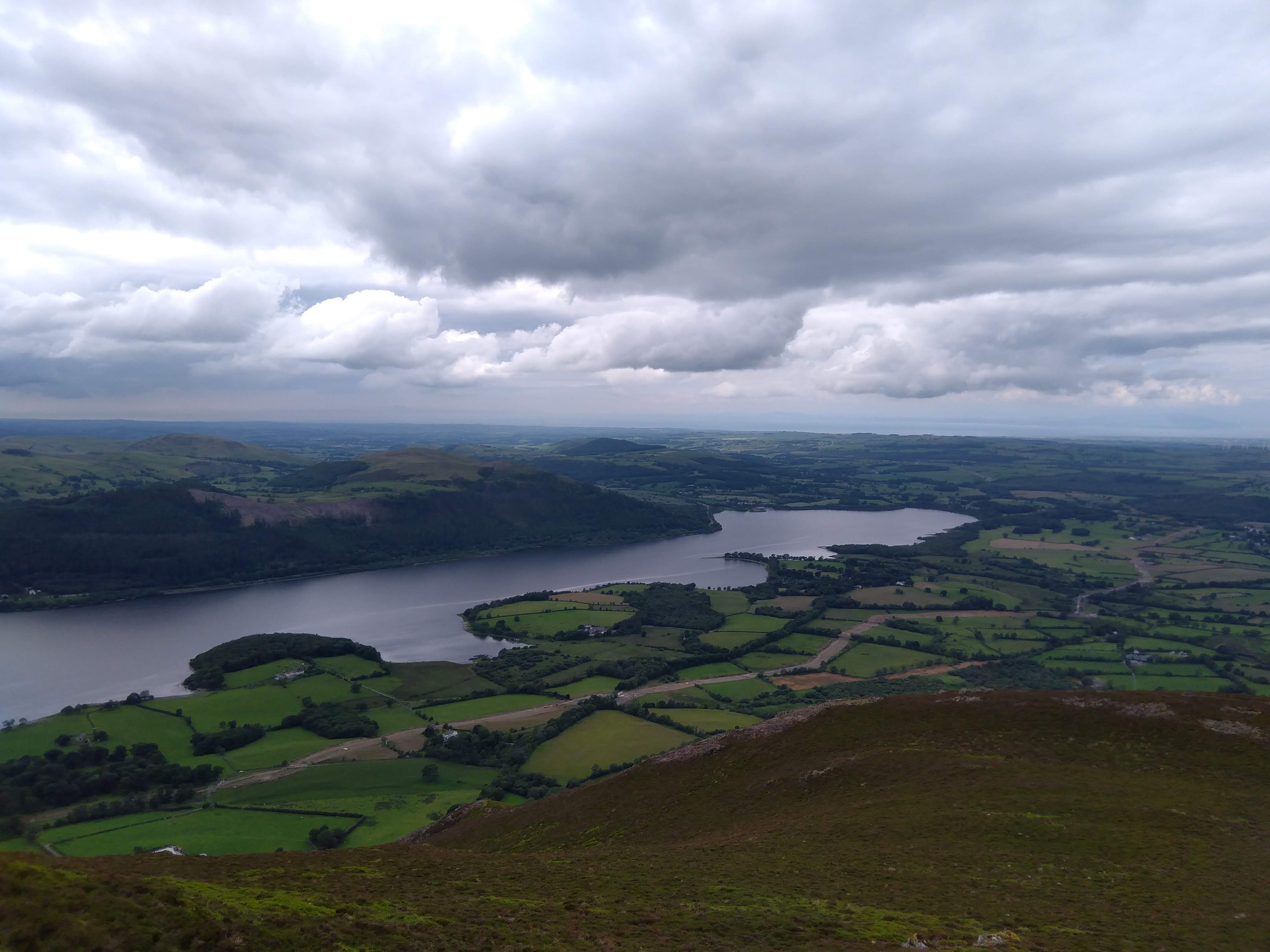 A sweeping, high-altitude landscape photograph taken from the slopes of Skiddaw mountain in the Lake District. The expansive view looks far down over a large lake, surrounded by a vibrant patchwork of green farm fields and rolling fells under a vast, dramatically cloudy sky.