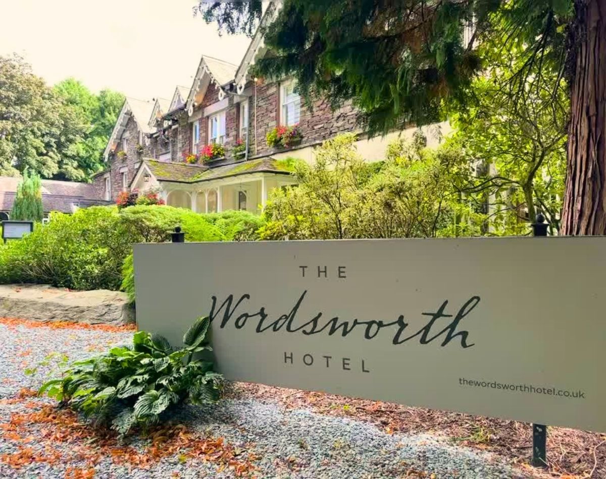 Elegant grey entrance sign for The Wordsworth Hotel sitting in front of the traditional Lakeland stone building and lush gardens in Grasmere village.