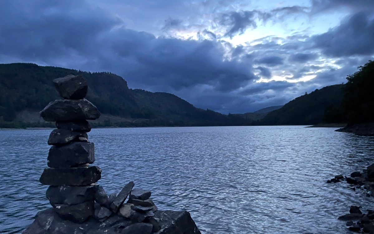 A balanced stack of stones sits on the rocky shoreline of Thirlmere reservoir against a dramatic blue dusk sky and dark silhouette of the fells.