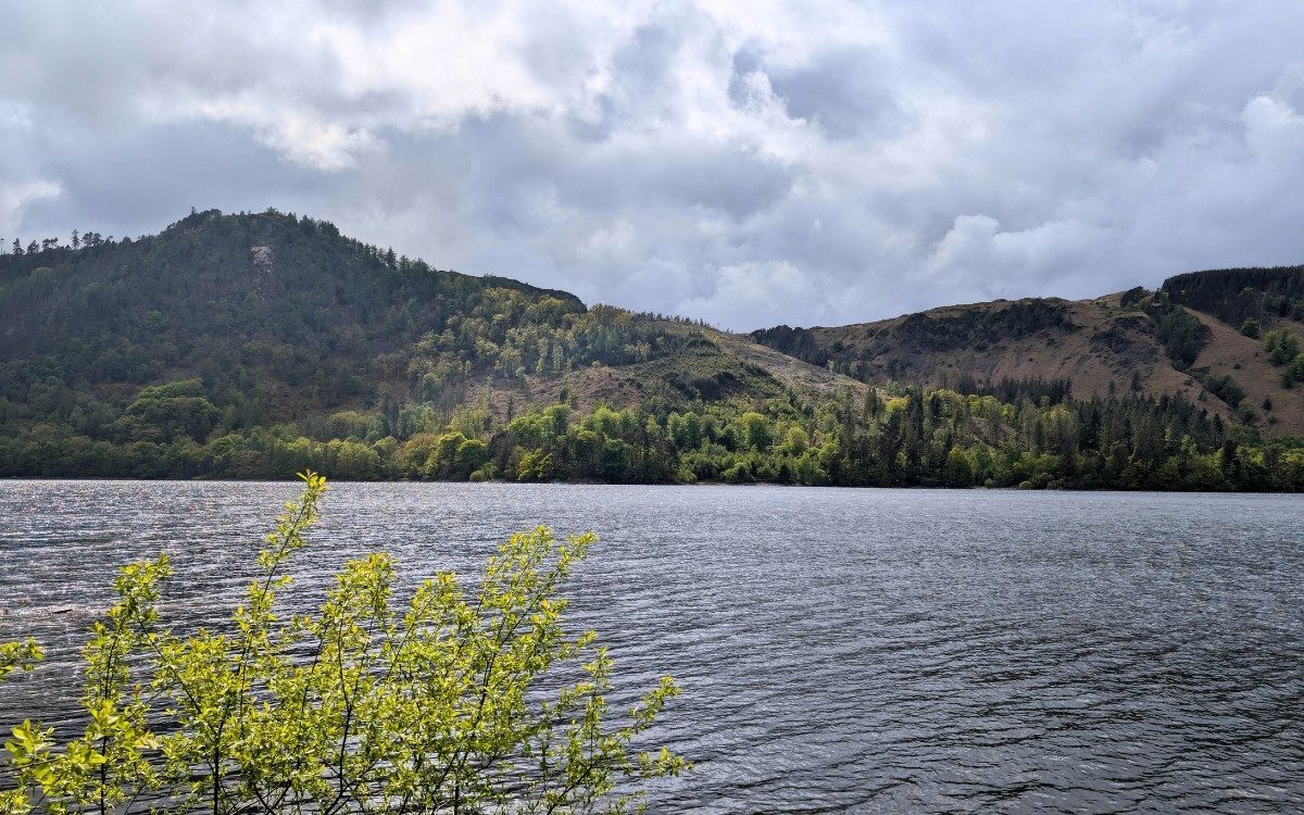 cenic daytime view across the rippling waters of Thirlmere reservoir towards forested fell slopes beneath a cloudy Lake District sky.