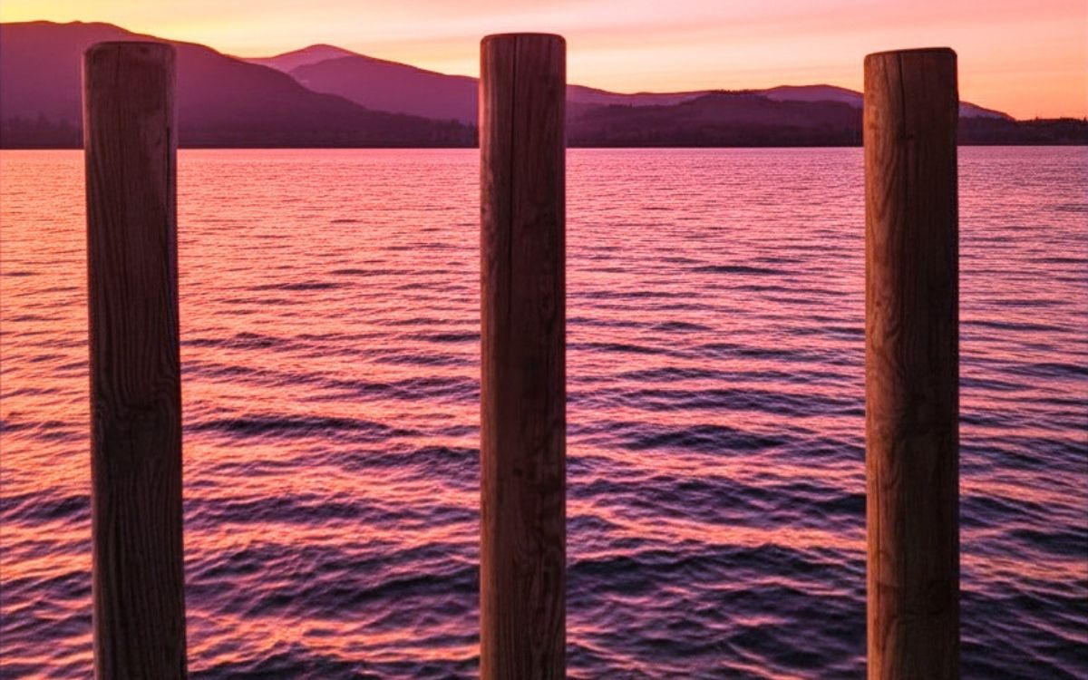 A vibrant pink and purple sunset reflecting on the calm waters of Derwent Water, viewed directly through the vertical wooden posts of Ashness Jetty in the Lake District.