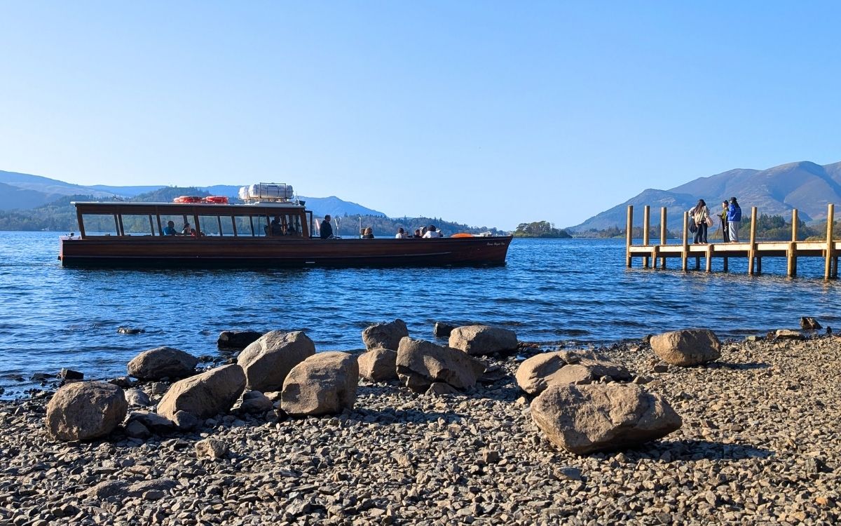 A classic wooden pier known as Ashness Jetty extending into the blue waters of Derwentwater, with a traditional wooden Keswick Launch about to pick up people.