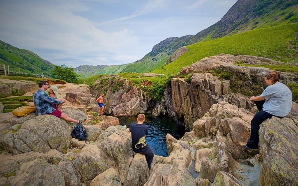 Crystal clear mountain pool for wild swimming in Borrowdale. A group of people relaxing on the craggy rock ledges surrounding the deep, dark plunge pool at Black Moss Pot in the Langstrath Valley.