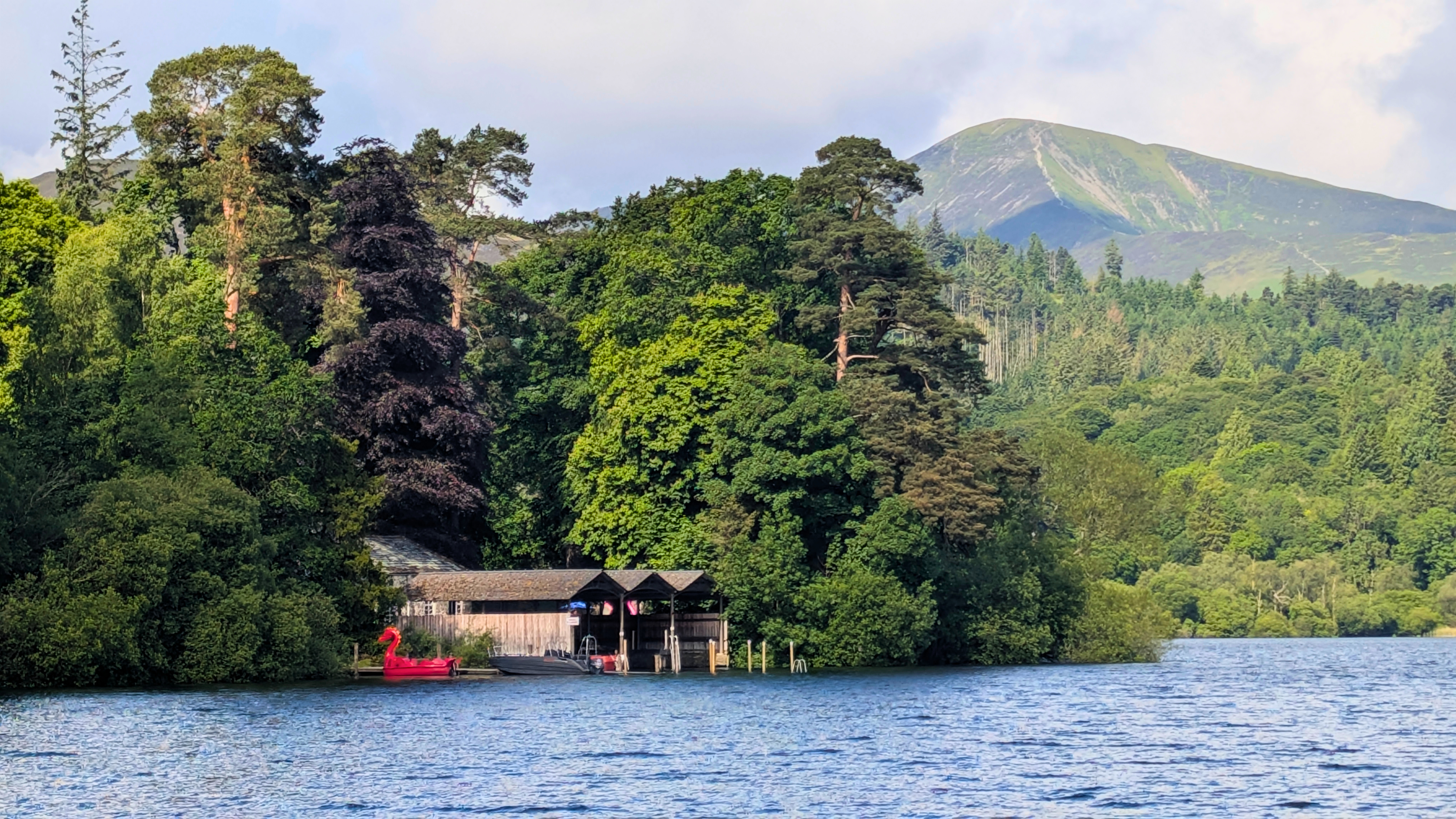 A crisp view across the rippling blue waters of Derwentwater towards the private Derwent Island. Traditional wooden boathouses are nestled among lush green trees on the water's edge, with a bright red dragon pedalo moored in front and a mountain peak rising in the background.