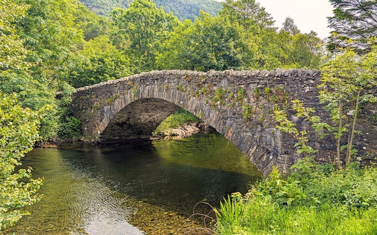 A classic double-arched stone bridge known as Grange Bridge crossing the clear, shallow waters of the River Derwent in Borrowdale, surrounded by lush green summer trees.