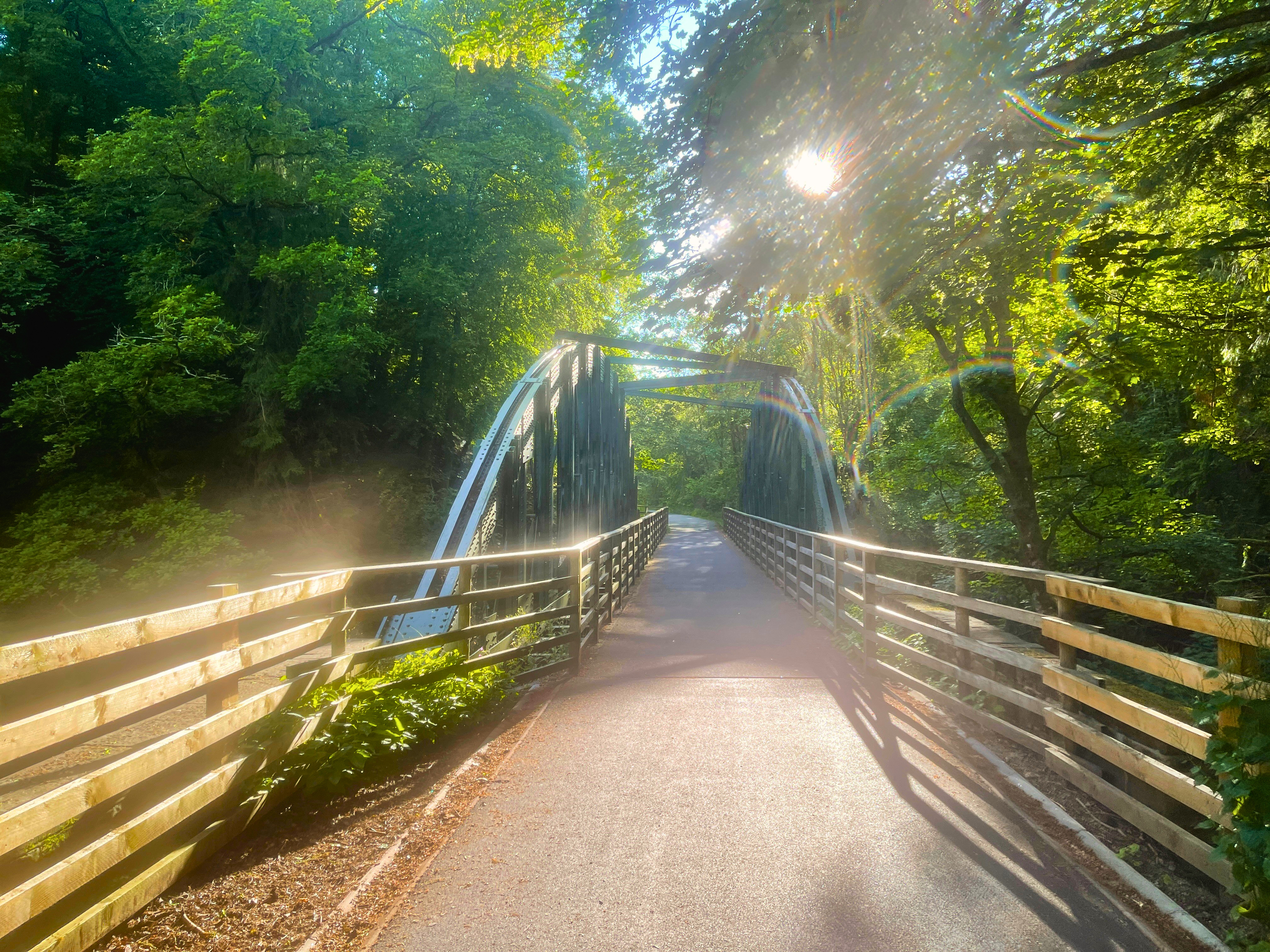 A smooth paved path with wooden fences leading to a large, historic dark green metal truss railway bridge on the Keswick to Threlkeld trail. Bright sunlight bursts through the lush green trees, creating a lens flare and a warm, hazy glow.