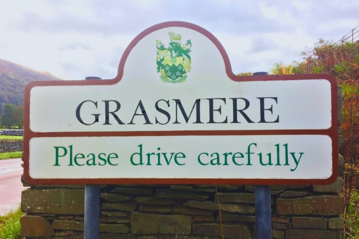 A decorative wooden welcome sign for Grasmere village, framed by vibrant green summer foliage.
