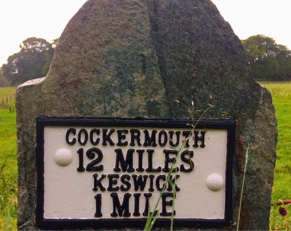 A classic white historic cast iron mile marker stone showing Cockermouth 12 miles and Keswick 1 mile, situated in a layby near Portinscale village.
