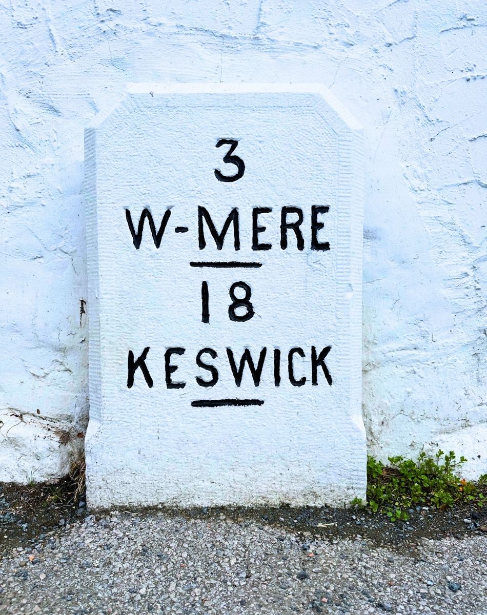 An ancient white milestone marker at Low Wood Bay showing 3 miles to Windermere and 18 miles to Keswick, set against a dark stone wall on the roadside.