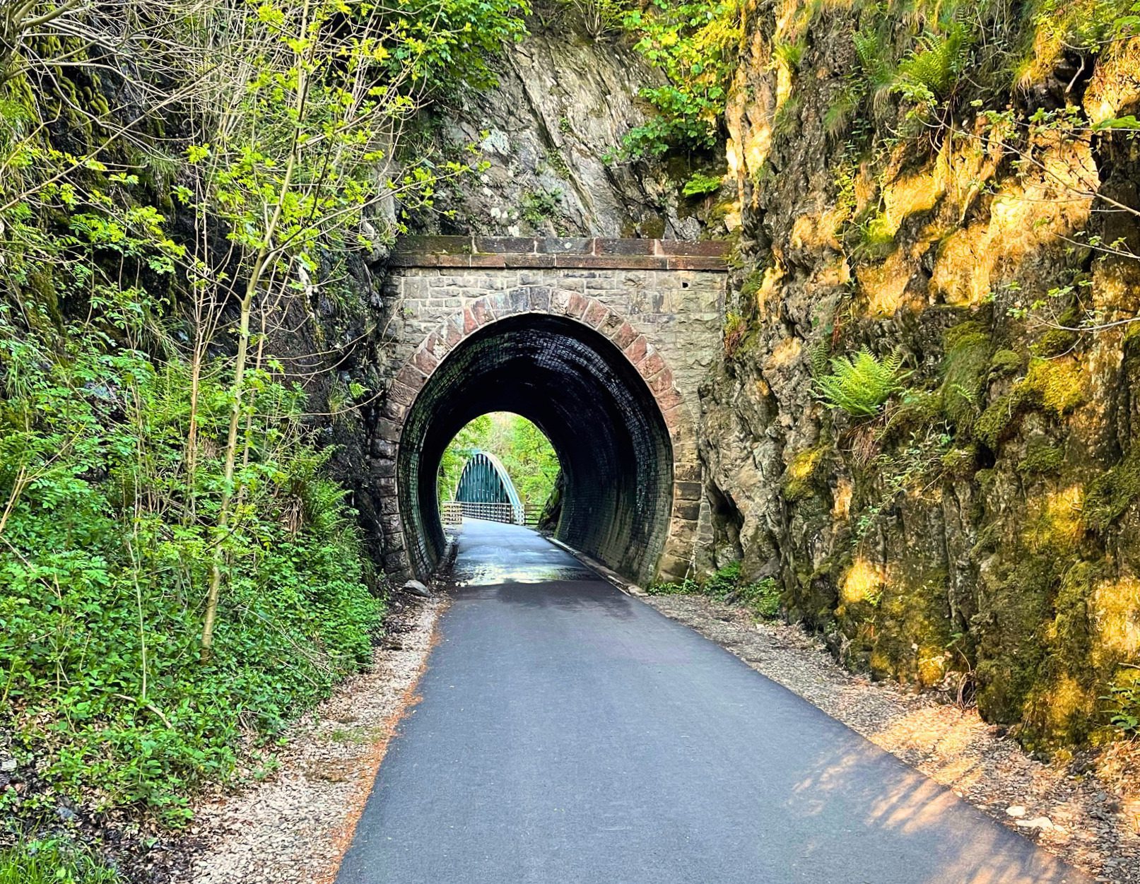 A smooth paved walking path leading into a historic stone arch railway tunnel on the Keswick to Threlkeld trail. The tunnel entrance is cut deep into rugged rock walls covered in lush green ferns and moss, with bright sunlight shining at the far end.
