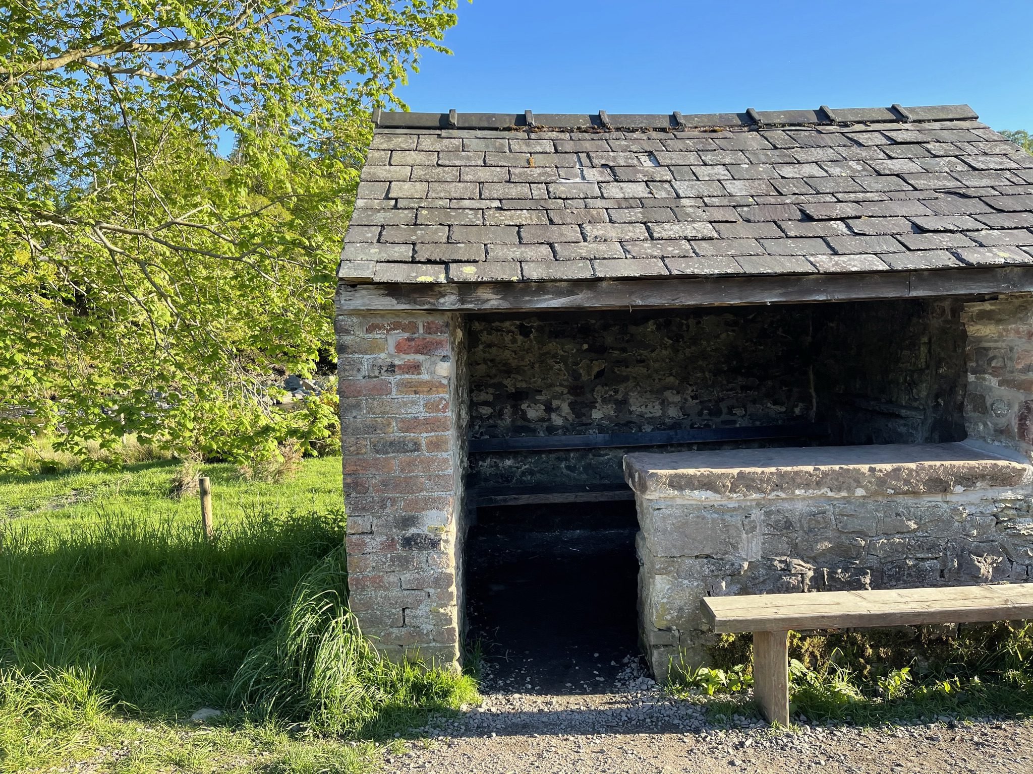 A rustic, historic brick and stone railway shelter with a traditional slate roof, known locally as the first hut, sitting beside the Keswick to Threlkeld trail next to vibrant green trees under a clear blue sky.