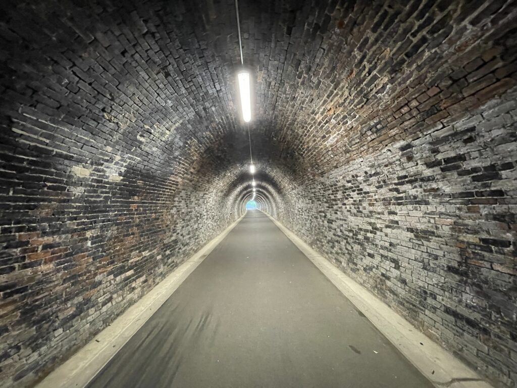 Looking down the center of a long, illuminated historic railway tunnel on the Keswick to Threlkeld trail. The tunnel features original arched brickwork and a smooth, accessible paved walking path.