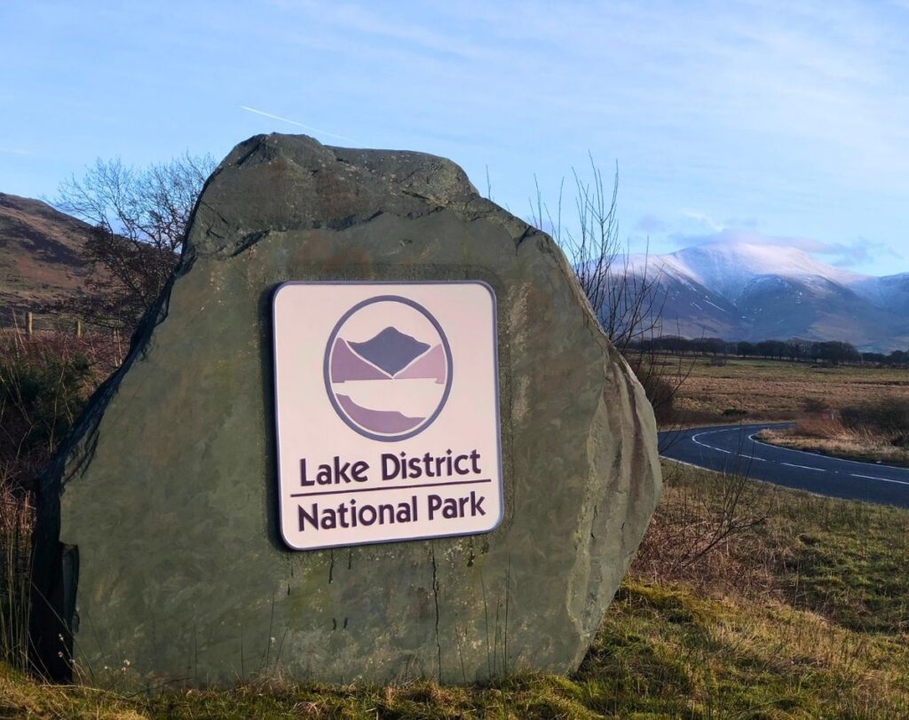 The brown Lake District National Park welcome boundary sign on the A591 road from Bothel, with the massive, snow-capped peaks of Skiddaw rising in the background.