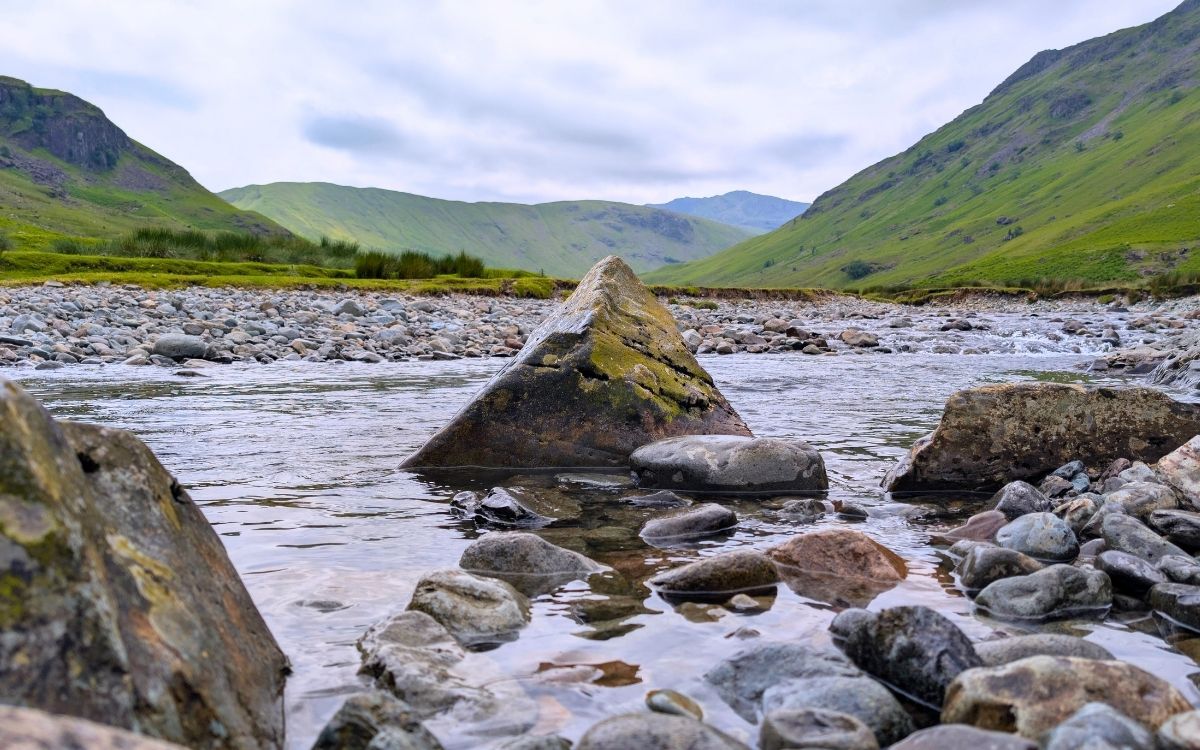 Low-angle view of clear water flowing around a large, mossy triangular rock and over a pebbly riverbed in Langstrath Beck, surrounded by the green fells of the Lake District.
