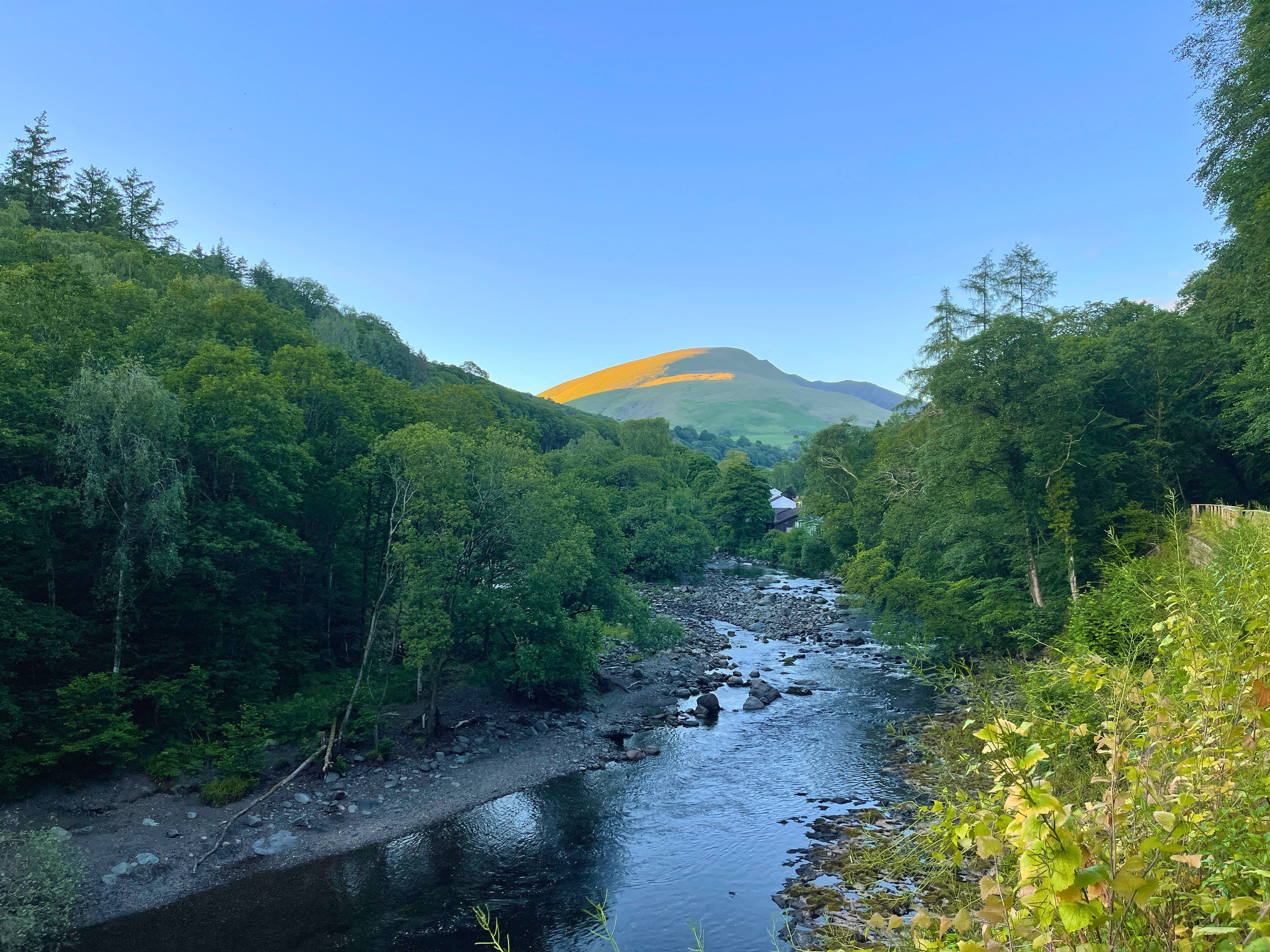 A scenic view looking up the rocky River Greta as it winds through a lush green wooded gorge, with the smooth, sun-kissed peak of Blencathra mountain standing prominently against a clear blue sky on the Keswick to Threlkeld trail.