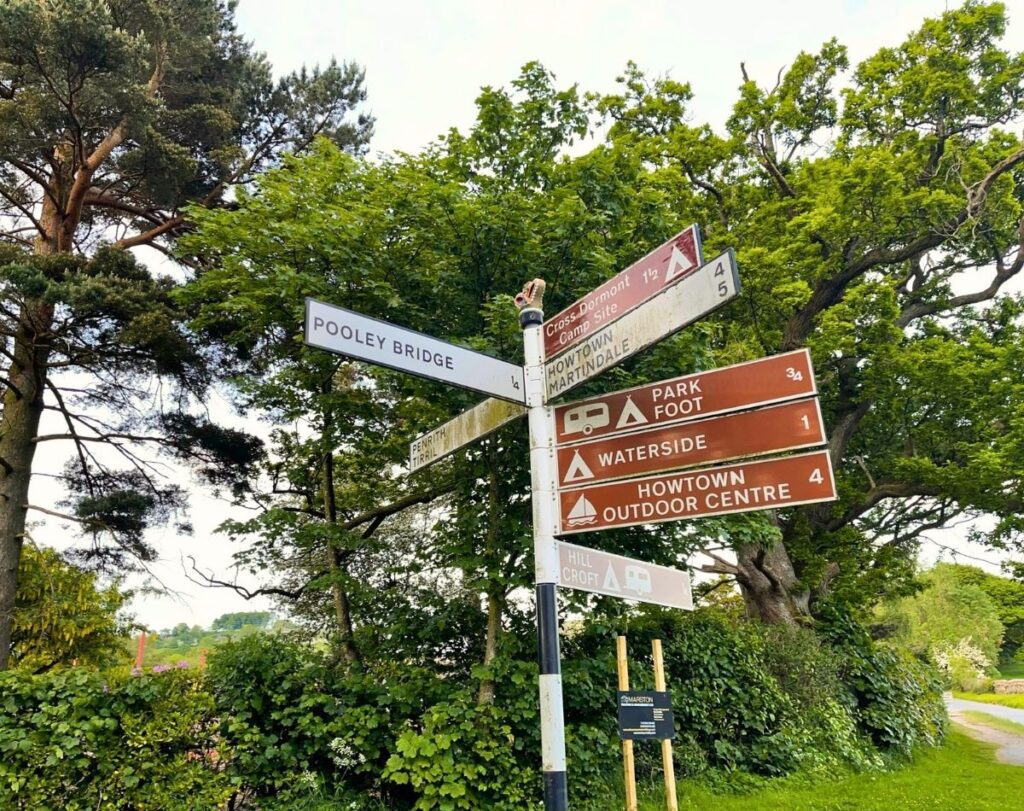 A traditional white metal directional signpost at a crossroads in Pooley Bridge, pointing towards local routes and village amenities.