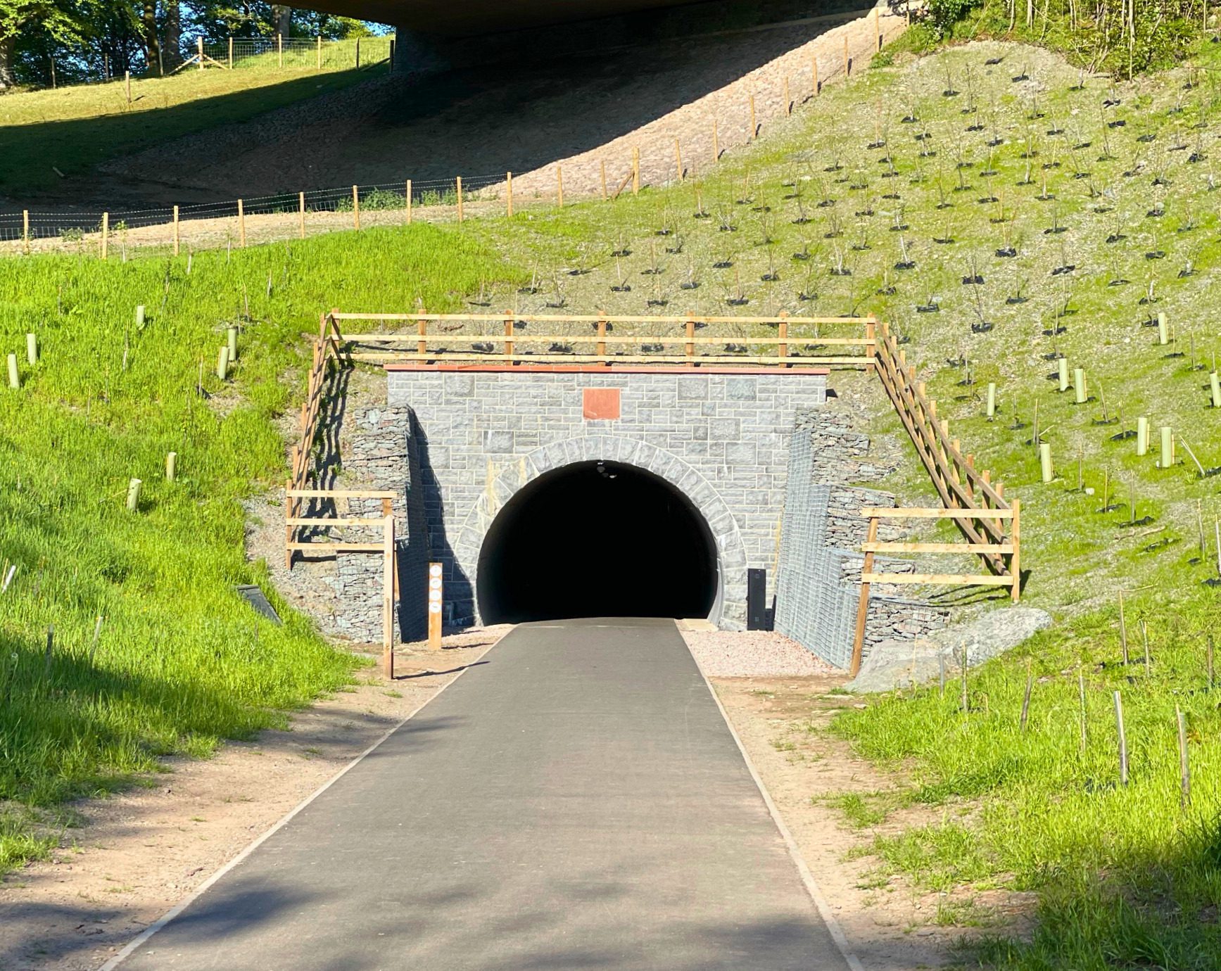A smooth, paved walking path leading into a beautifully restored stone-faced railway tunnel on the Keswick to Threlkeld trail. The entrance is framed by wooden fencing, and grassy embankments under a bright sky.