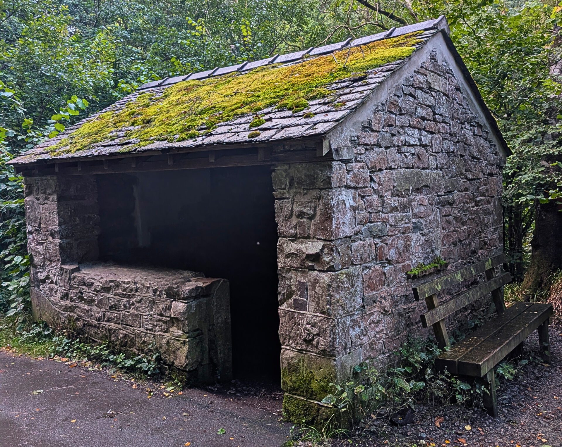 A traditional, rustic stone railway shelter featuring a slate roof covered in bright green moss, known locally as the second hut on the Keswick to Threlkeld trail. A wooden bench sits outside on a damp, leaf-strewn paved path surrounded by dense woodland.