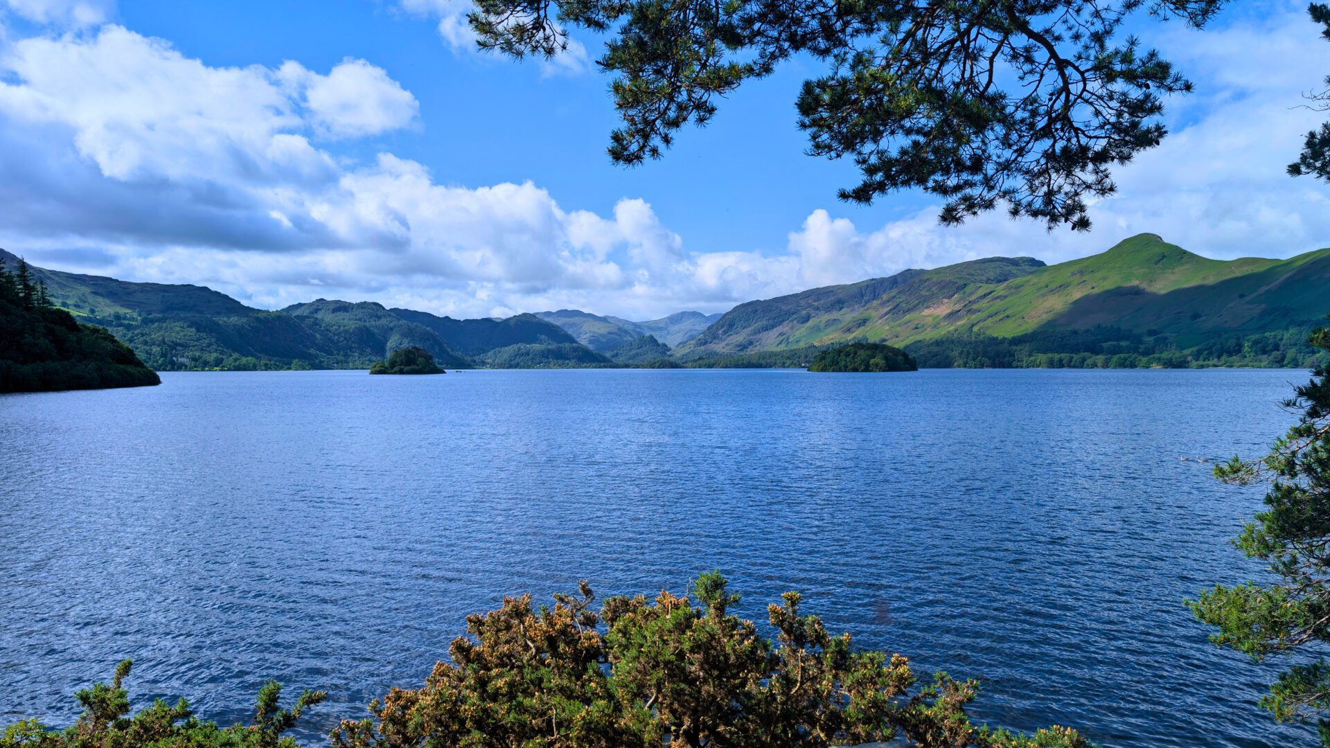 Southern View from Friars Crag over Derwentwater Islands Alt Text: A stunning landscape view looking south from Friars Crag over the vibrant blue waters of Derwent Water. Both St Herbert's Island and Lord's Island are clearly visible, surrounded by dramatic Cumbrian mountains under a bright, partly cloudy sky.