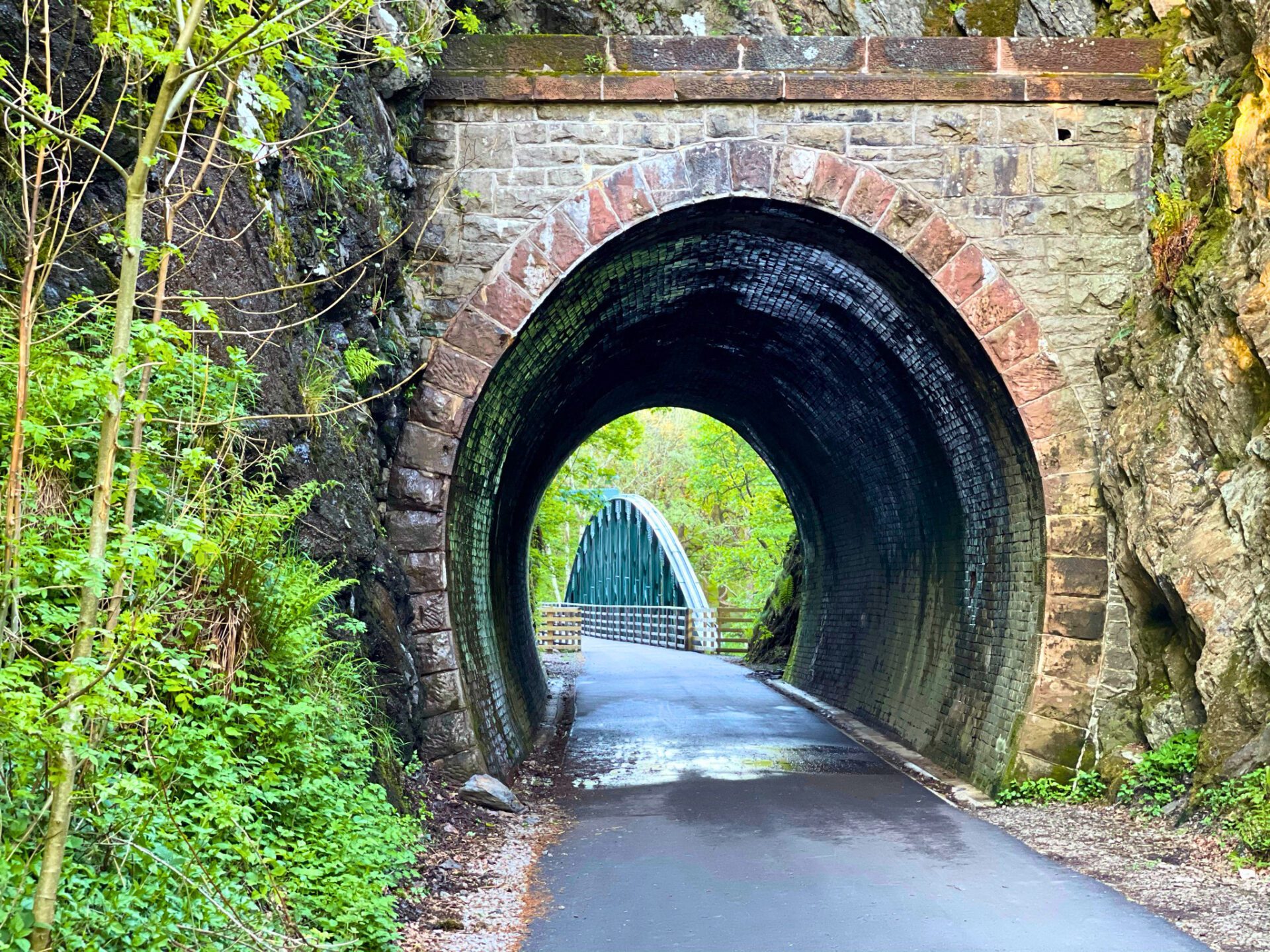 A damp, paved walking path leading through a dark, historic stone arch railway tunnel on the Keswick to Threlkeld trail. Perfectly framed through the tunnel's exit is the iconic, dark green metal railway bridge surrounded by bright summer foliage.