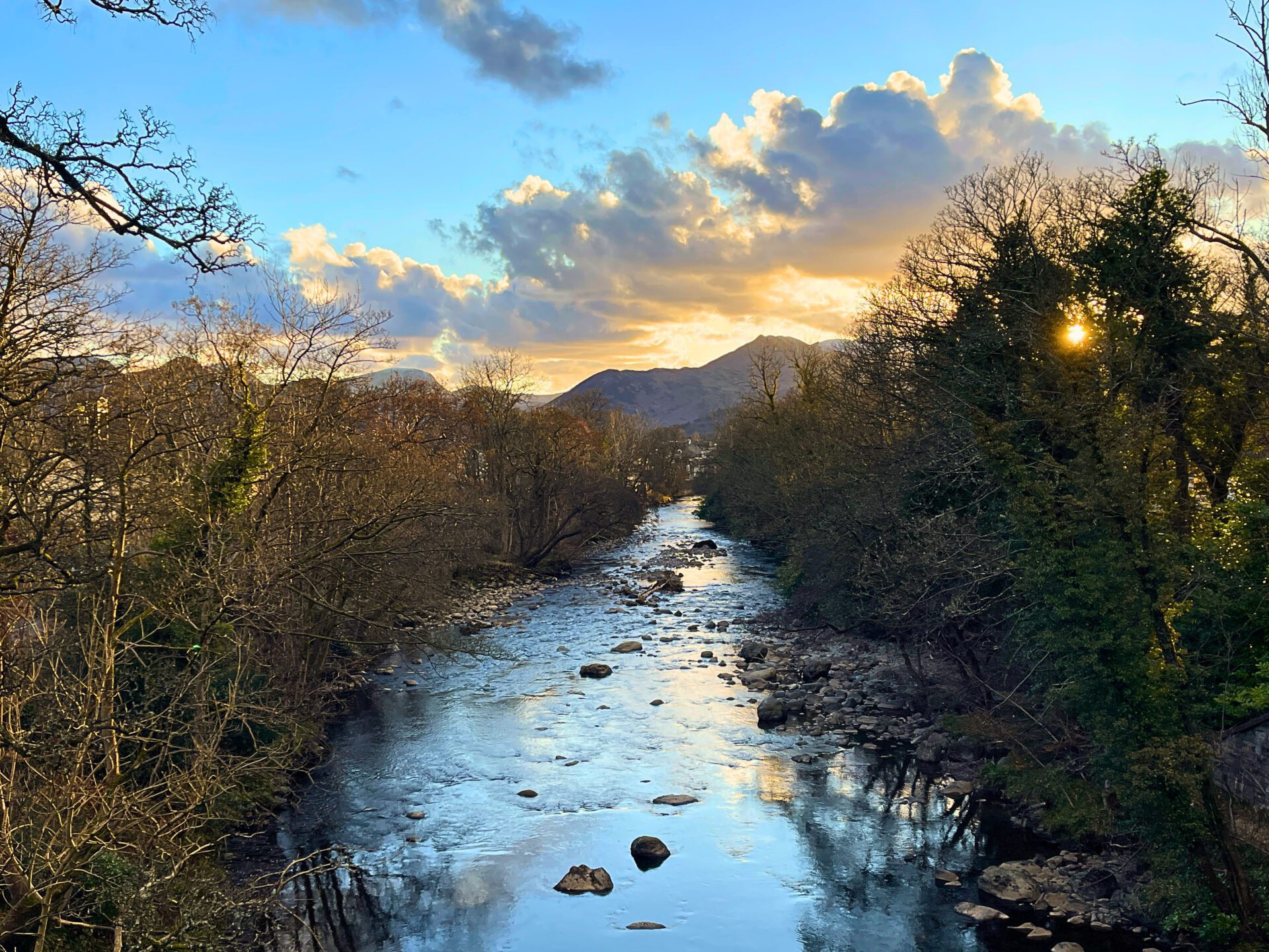 A scenic winter view looking down the rocky River Greta towards Keswick town from the first bridge on the trail, framed by bare trees with the sun peeking through and distant mountains under a dramatic cloudy sky.