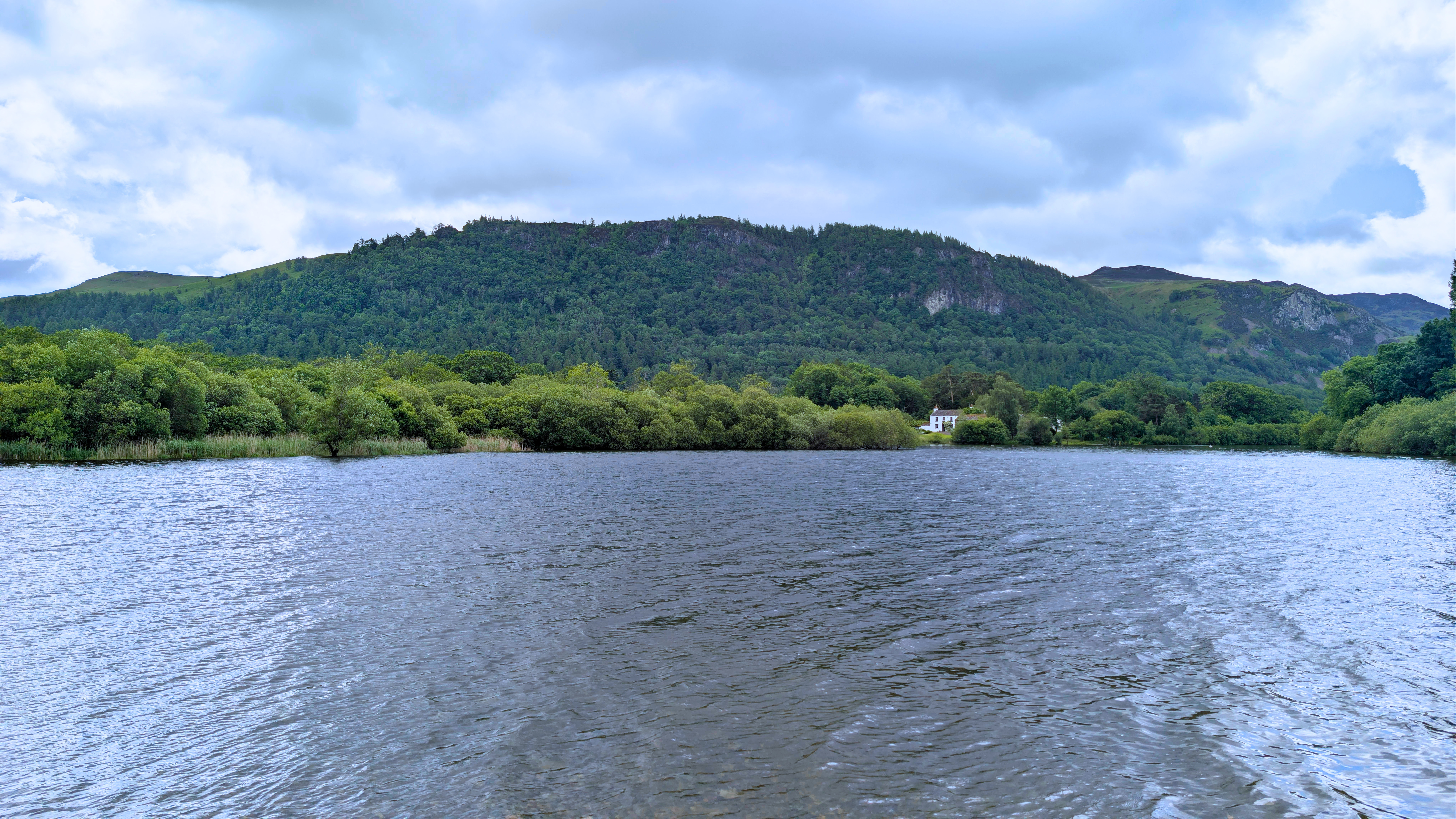 A wide, scenic view across the rippling waters of Strandshag Bay on Derwentwater, looking towards a lush, tree-lined shore and the prominent, forested heights of Walla Crag under a bright, cloudy sky.