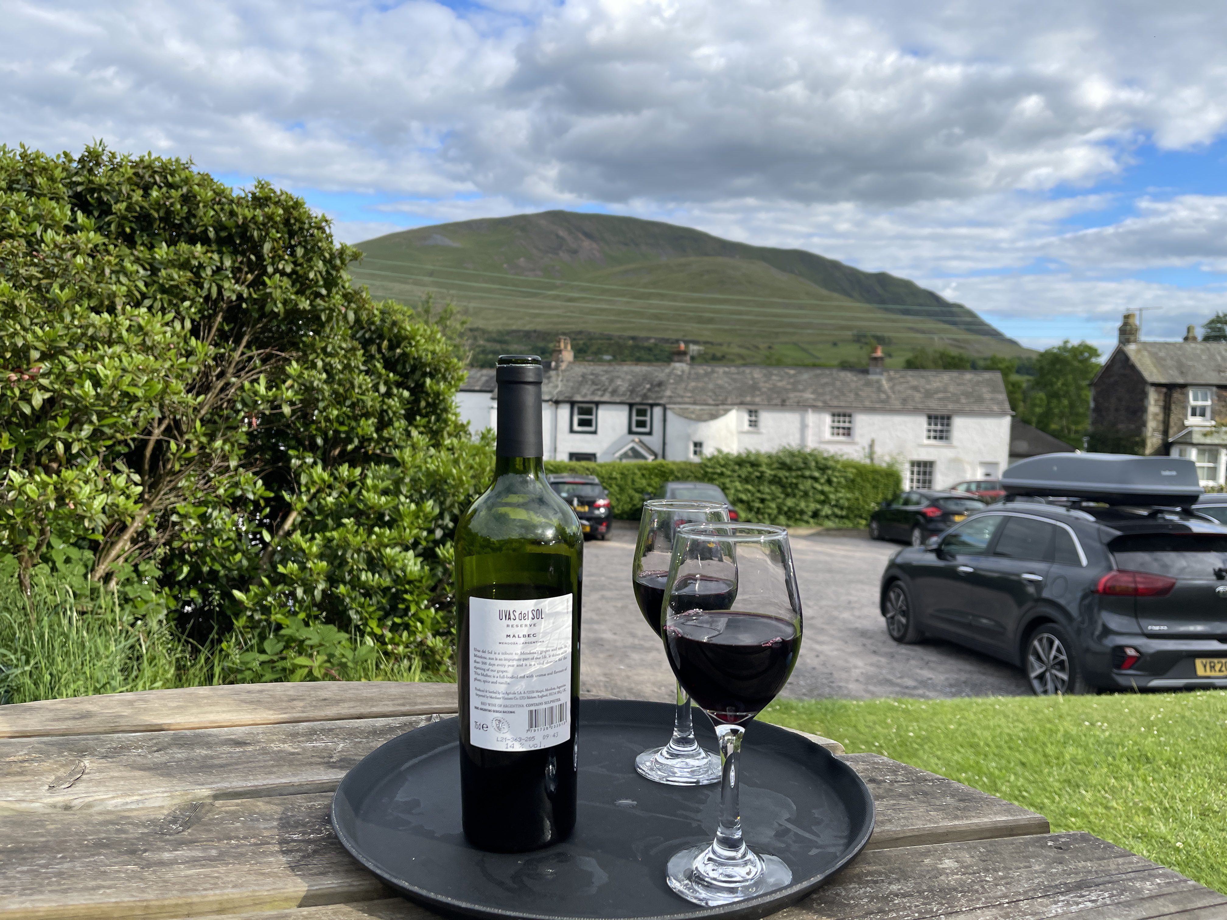 A bottle of red wine and two filled glasses resting on a black tray on a wooden picnic table in the Horse and Farrier beer garden in Threlkeld. In the background sits the traditional white pub, a car park, and the sweeping green slopes of the fells under a cloudy blue sky.