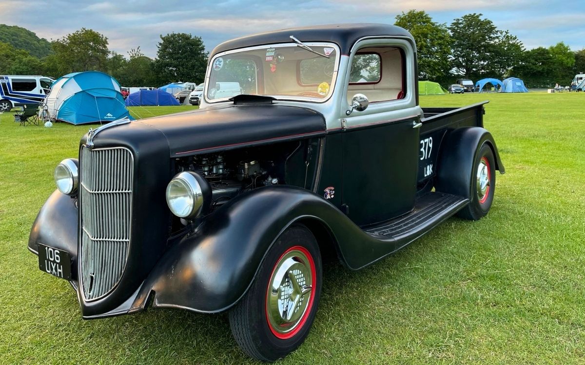 Custom matte black classic hot rod pickup truck with chrome grille and red steel wheels parked at a Lake District campsite.
