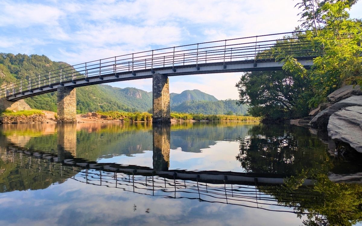 A low-angle view from the surface of the River Derwent, showing the stone pillars and metal span of the Chinese Bridge perfectly reflected in the calm water of the Borrowdale Valley.