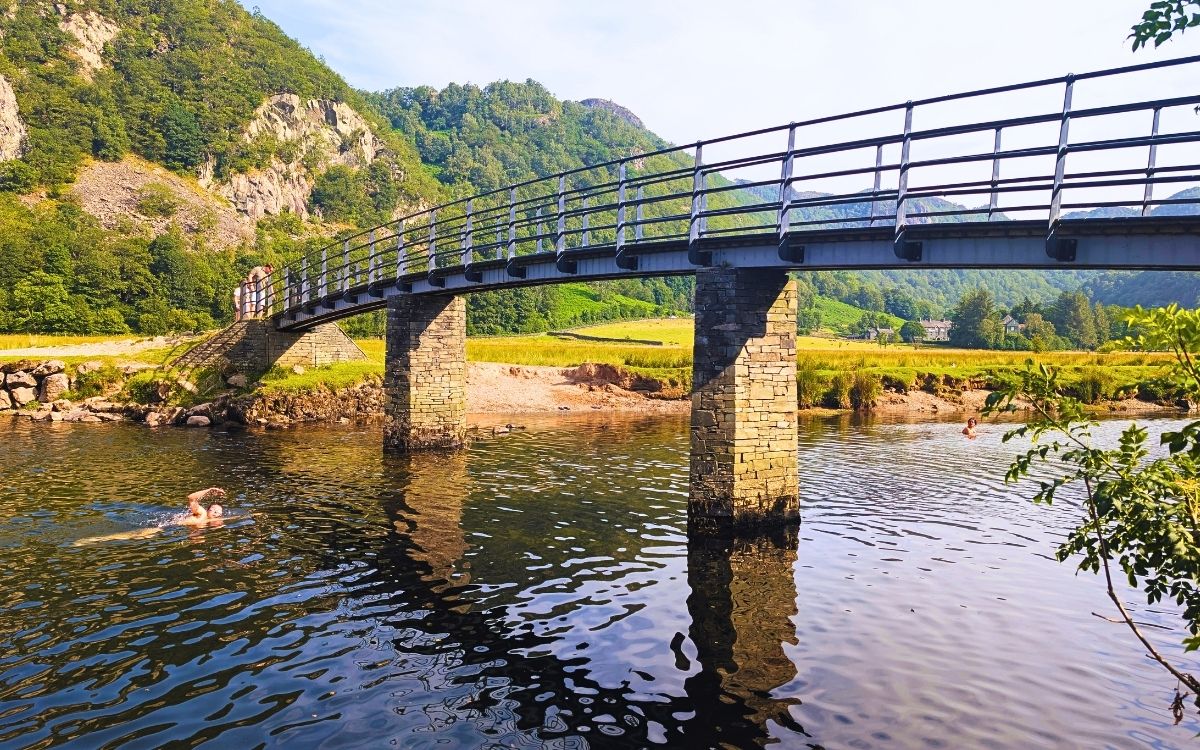 People wild swimming in the River Derwent beneath the stone pillars of the Chinese Bridge, a scenic crossing point on the Derwent Water walk in the Borrowdale Valley.