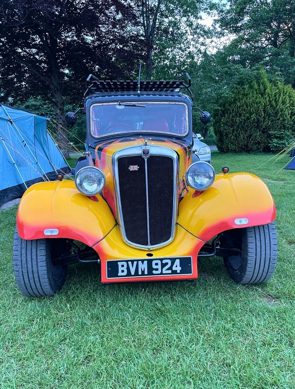 Straight front view of a custom modified classic British car featuring a vibrant yellow, orange, and red flame paint job on grass.
