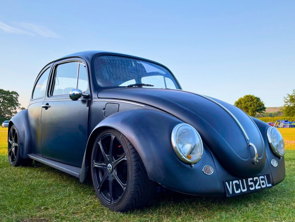 Customised matte black classic VW Beetle lowered on modern black alloy wheels parked on a grassy field at Parkfoot Campsite, Pooley Bridge.
