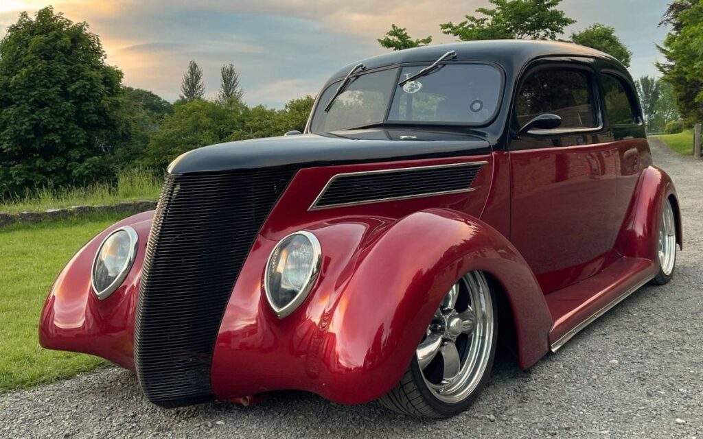 Custom slammed classic hot rod painted maroon and black with large chrome wheels parked on a gravel driveway at sunset.