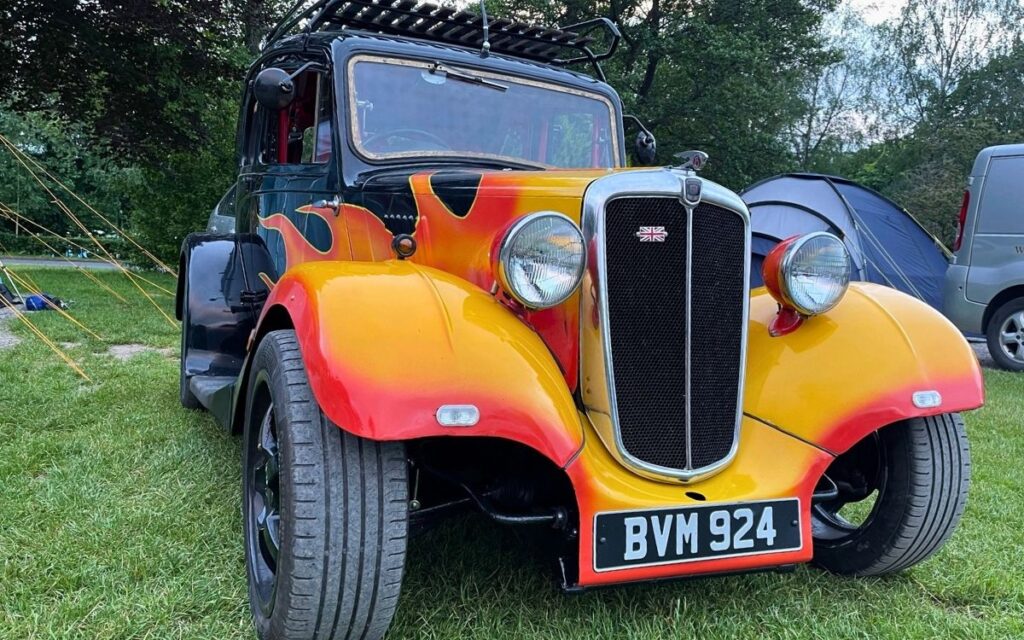 Three quarter close up view of a custom flame painted vintage car with roof rack parked next to tents at a Cumbrian campground.