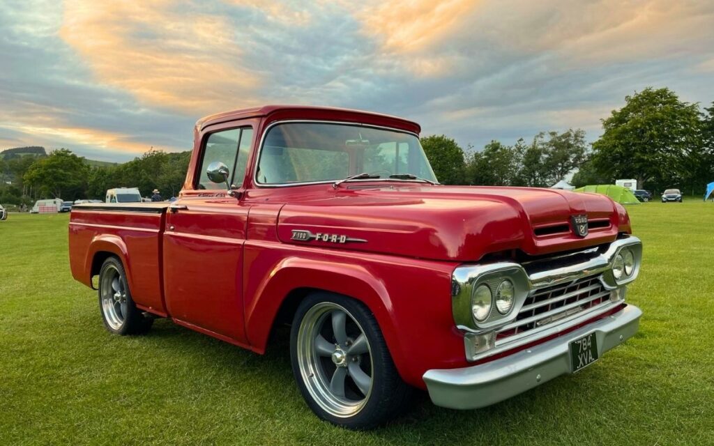 Restored glossy red classic Ford F100 pickup truck with custom wheels parked on a field under a sunset sky in the Lake District.