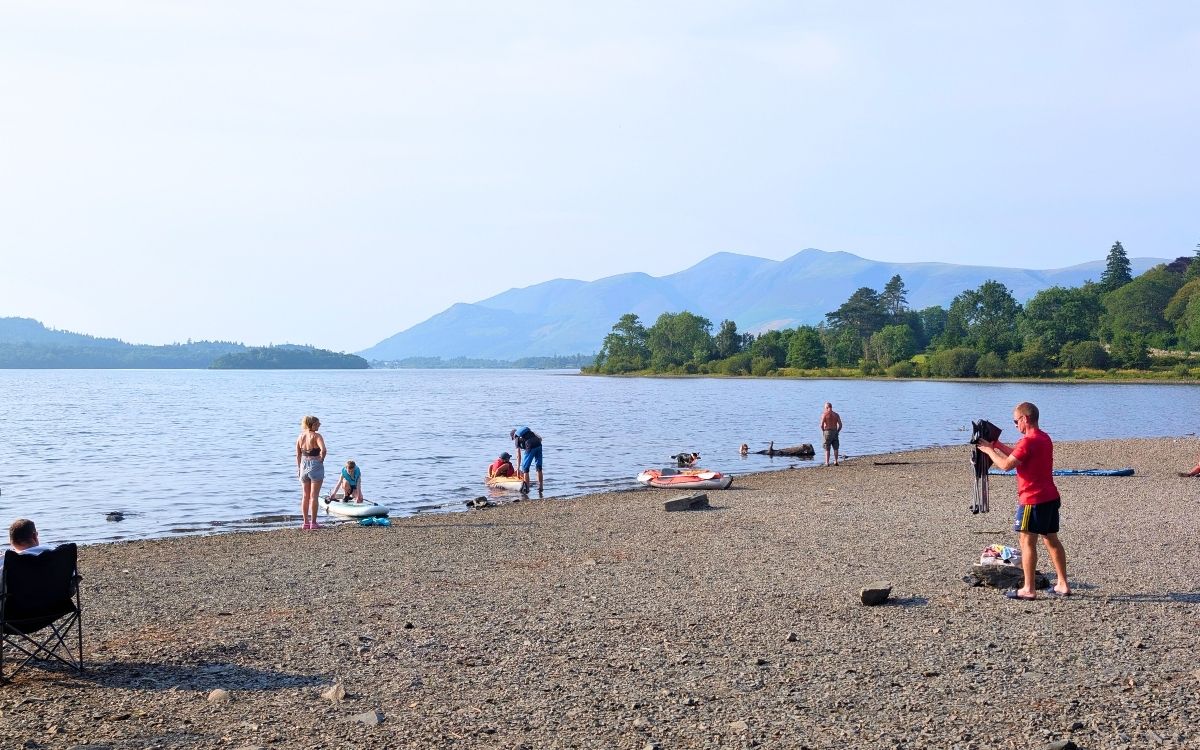 People enjoying a sunny day at the Kettlewell beach in Borrowdale, featuring swimmers, kayakers, and someone sitting in a camping chair looking out over Derwent Water towards Skiddaw.