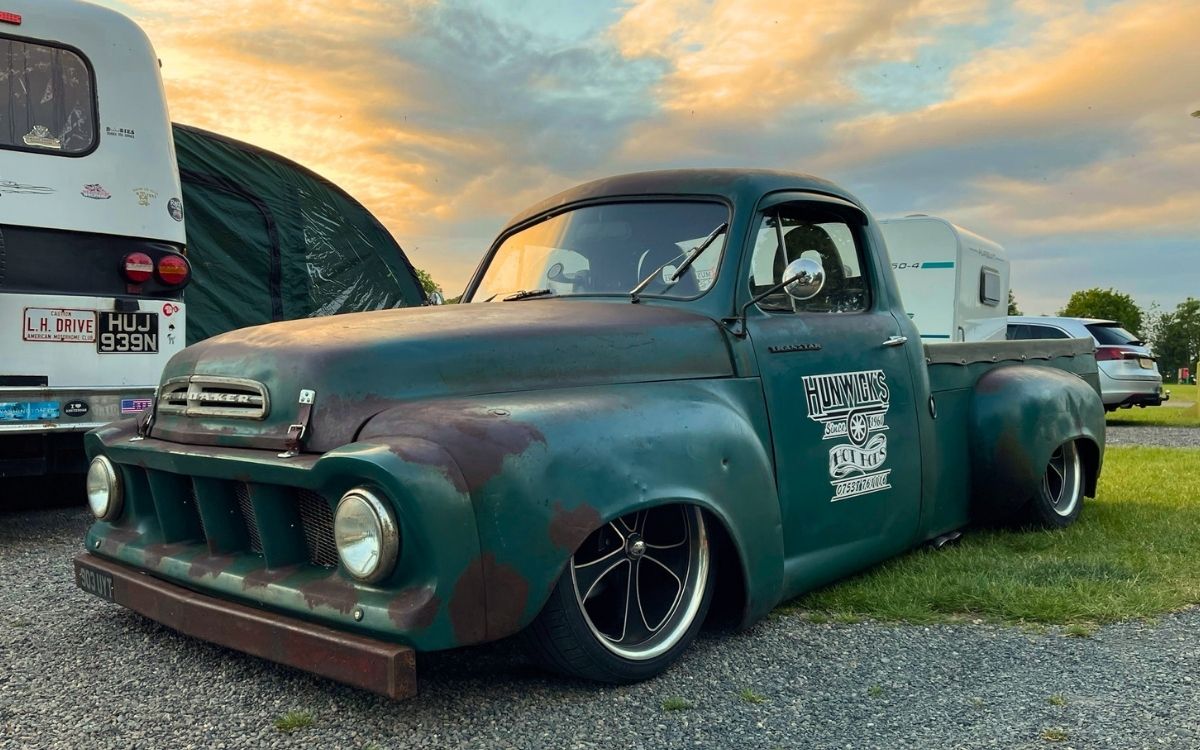 Lowered vintage green custom truck with Hunwicks Hot Rods sign writing parked on gravel during a Cumbrian sunset.