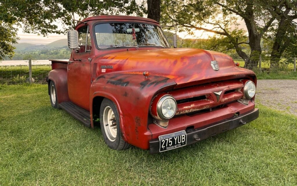 Custom lowered classic red Ford pickup truck with weathered patina paint parked on grass overlooking a Cumbrian lake and fells at Ullswater