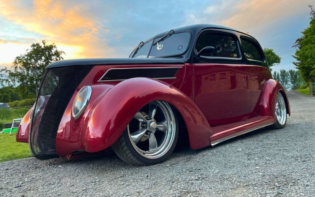 Custom slammed classic hot rod painted maroon and black with large chrome wheels parked on a gravel driveway at sunset.