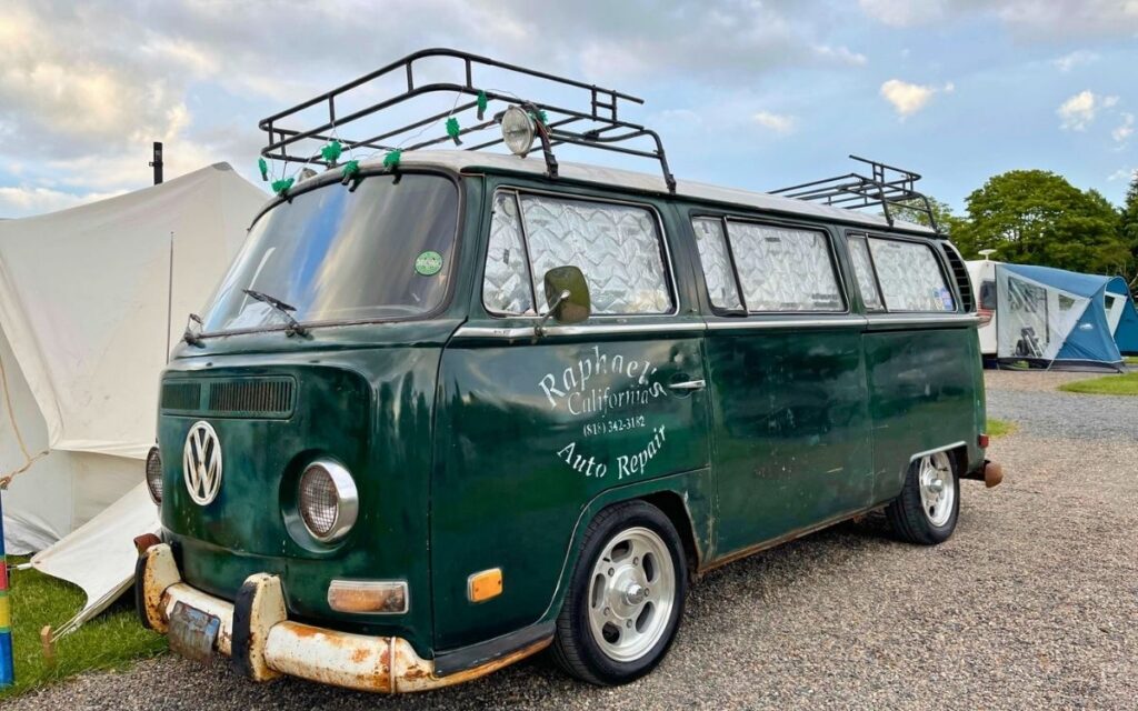 Classic dark green vintage VW campervan with California Auto Repair sign writing and a roof rack parked on a gravel campsite in the Lake District.
