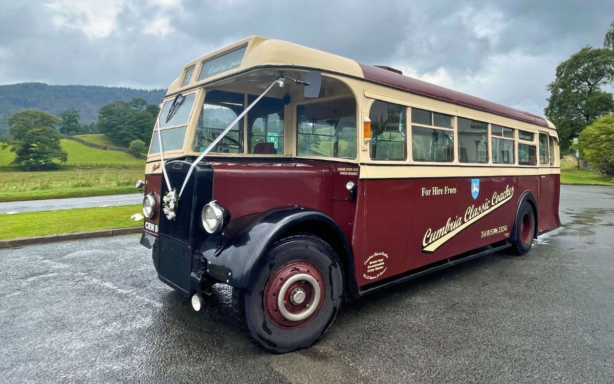 Maroon and cream vintage Cumbria Classic Coaches bus decorated with a white wedding ribbon parked against a backdrop of Lake District hills.
