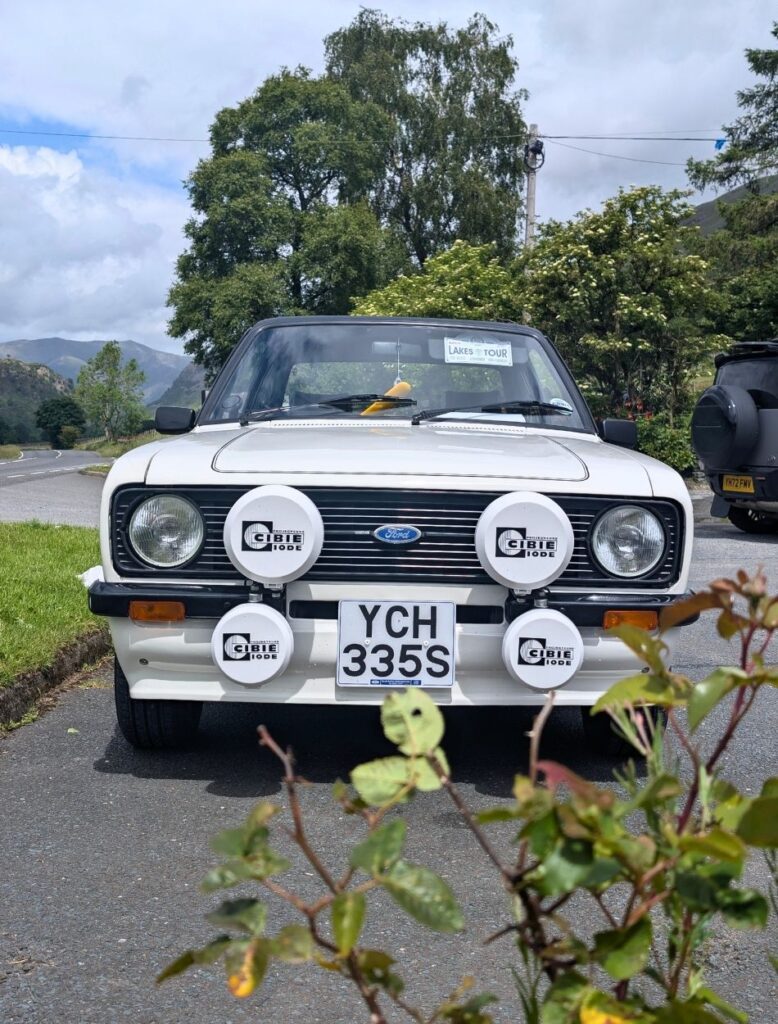 Front view of a classic white Ford Escort with vintage rally spotlights parked on a scenic road (A591) during a Lake District tour.