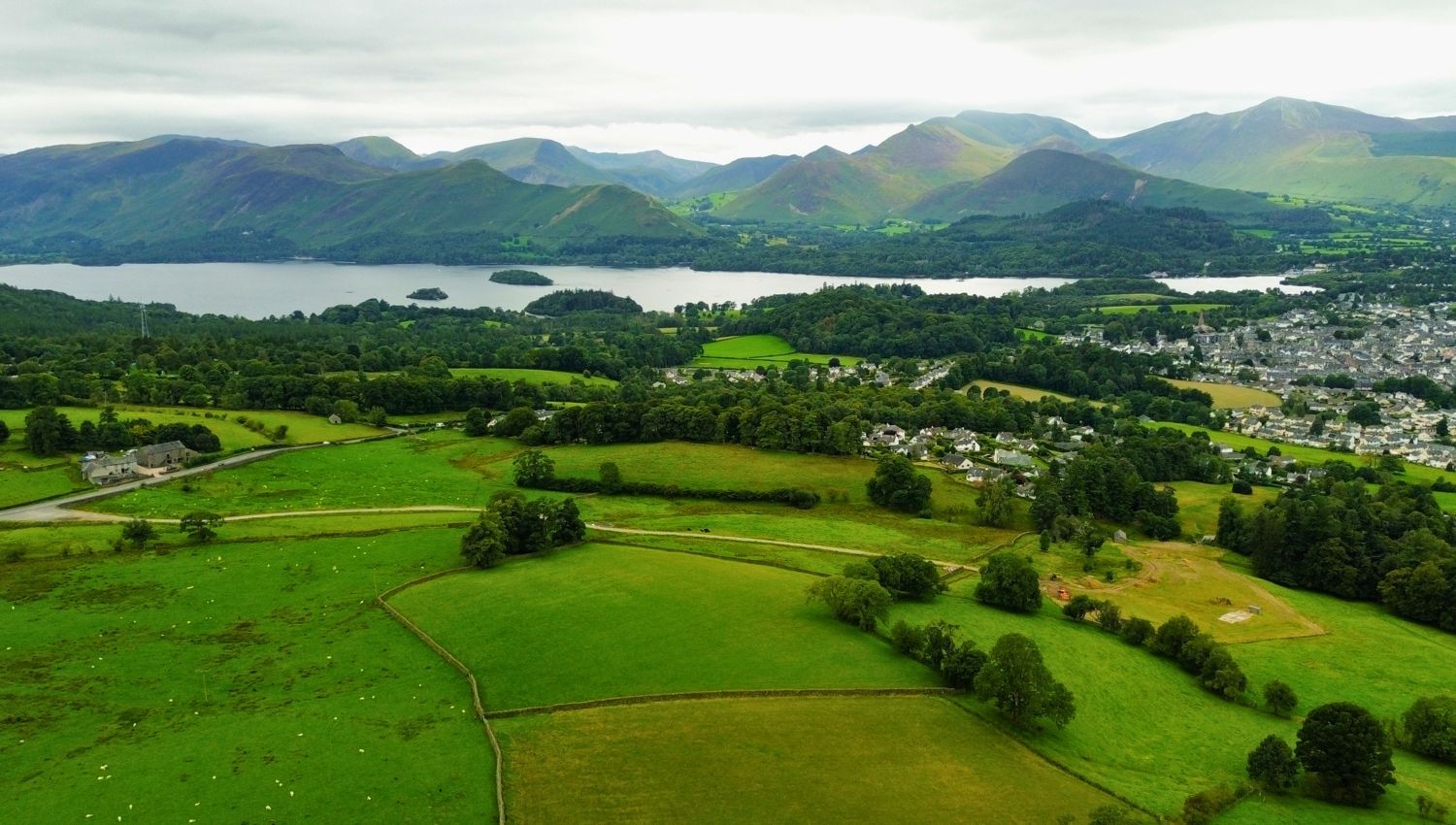 An expansive aerial drone shot taken from above Castlerigg looking out over the town of Keswick, Derwent Water, and the prominent peaks of Catbells and Grizedale Pike.
