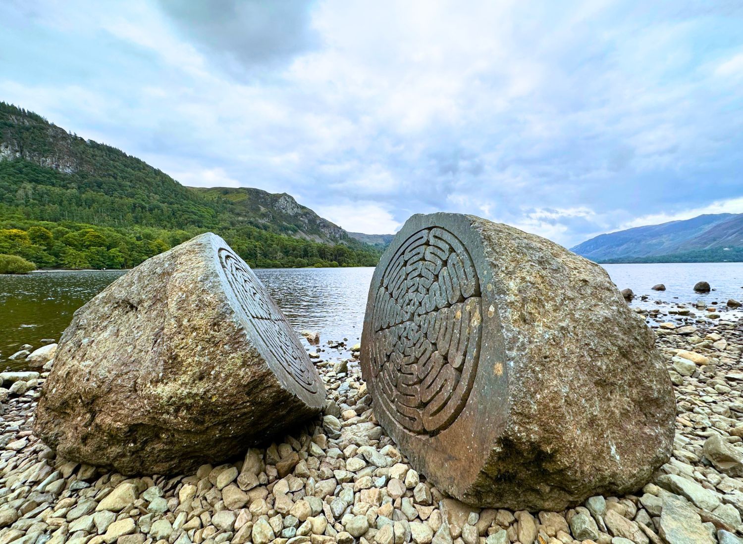 The National Trust Centenary Stone, a large split rock sculpture with intricate carved patterns on its inner faces, resting on the pebbled shore of Calfclose Bay at Derwent Water.