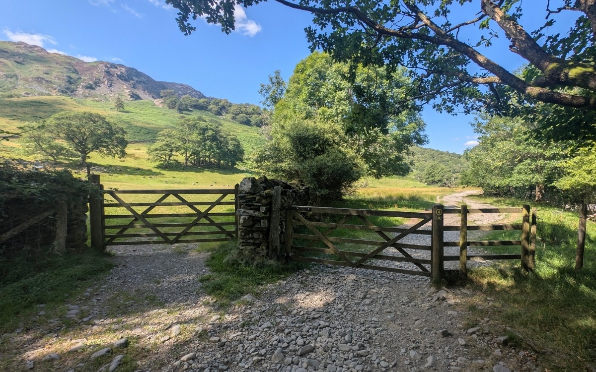 Pair of wooden farm gates across a stony path in Borrowdale, showing the right-hand gate with a yellow waymarker to continue along the track towards Castle Crag and the River Derwent.