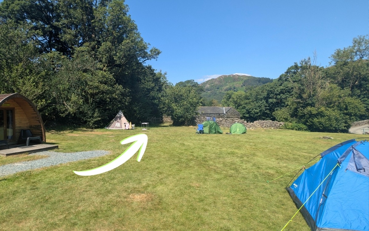 Camping field at YHA Borrowdale with glamping pods and tents, with a white arrow indicating the route towards the small wooden gate in the far left corner.
