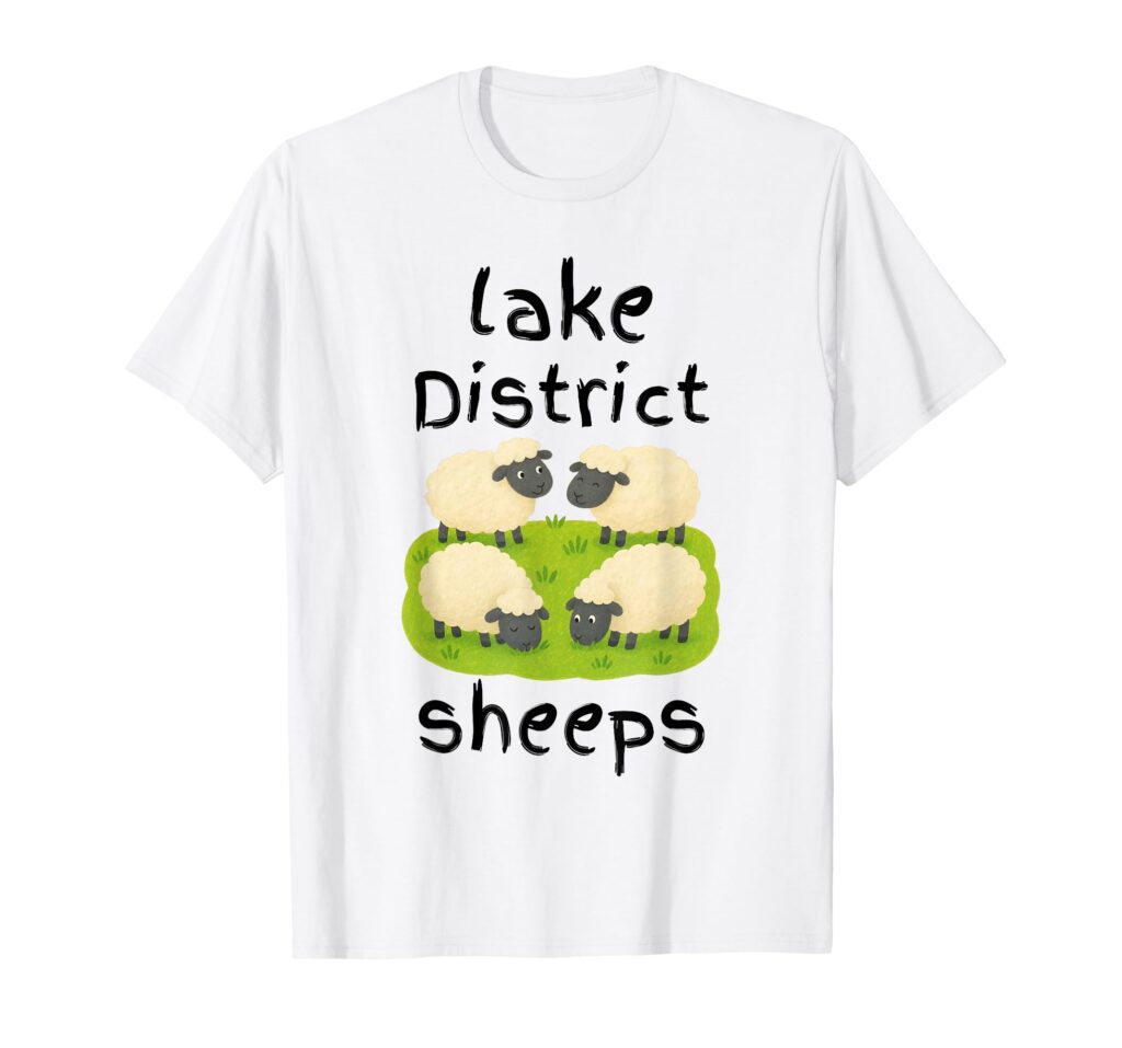 A white t-shirt featuring a cute cartoon illustration of four sheep grazing on a green patch of grass, with the text 'lake District' above and the playful, intentional misspelling 'sheeps' written below.