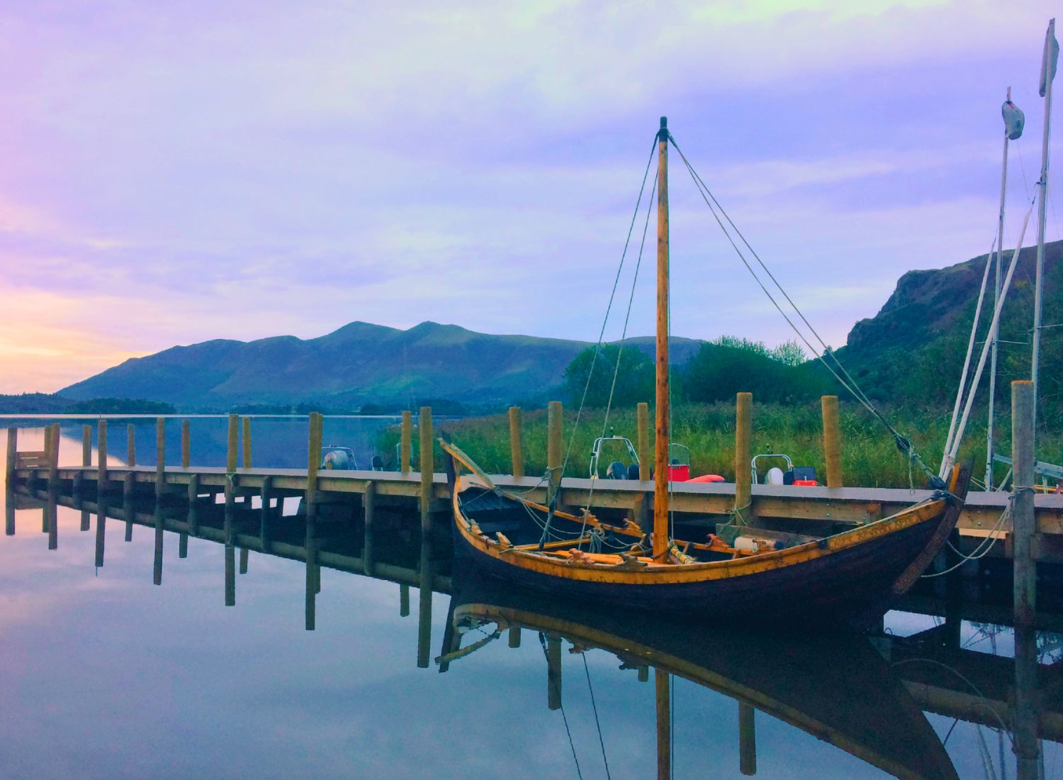 A traditional wooden boat moored at Lodore jetty in Borrowdale, floating on perfectly calm, reflective waters on Derwent Water with the Skiddaw mountain range in the background at dusk.